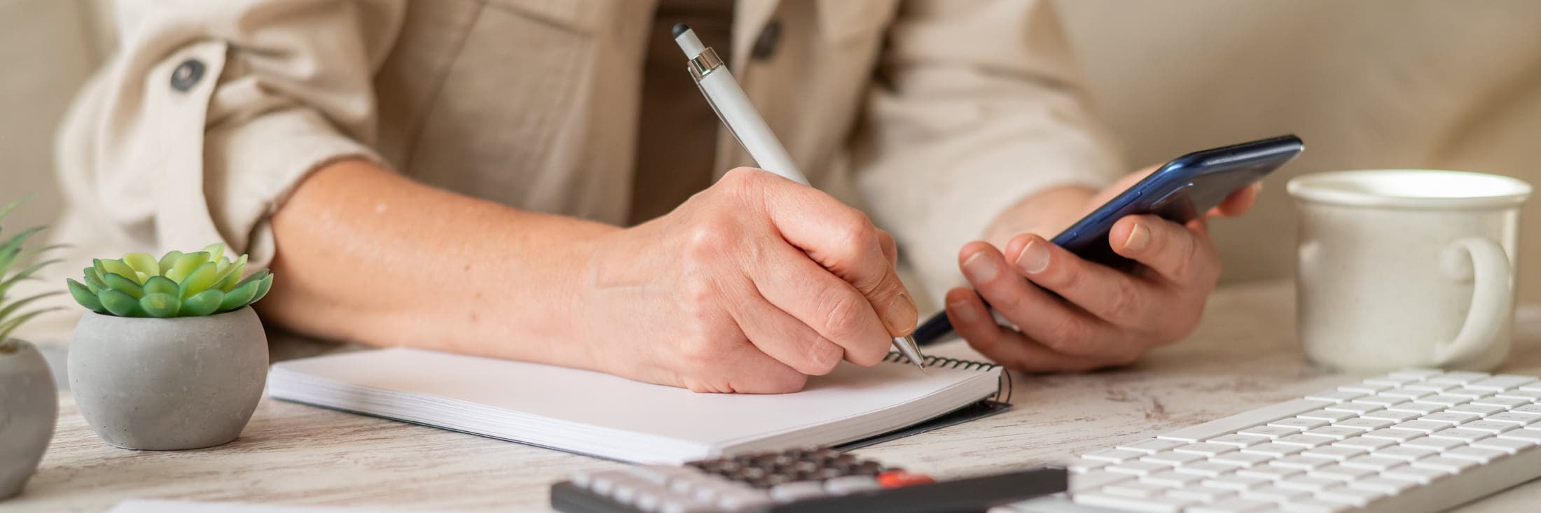A person's hands at a light table, writing in a spiral notebook with one hand and holding a phone in the other, a calculator and a keyboard beside them.